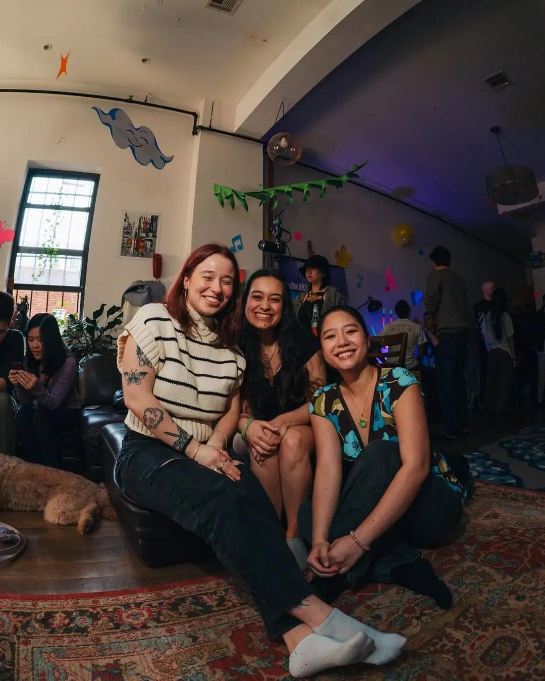 Three friends smiling and sitting together at a cozy indoor gathering with colorful wall decorations.