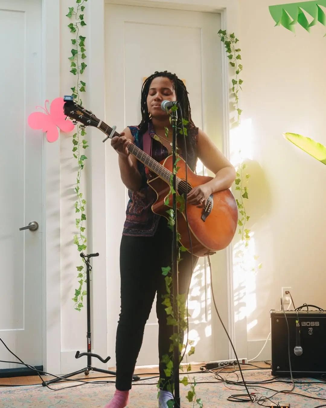 Musician performing with acoustic guitar and microphone, surrounded by leafy decorations indoors.