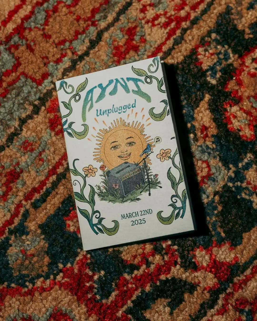 A decorative event flyer for 'AYNI Unplugged' placed on a colorful patterned carpet.