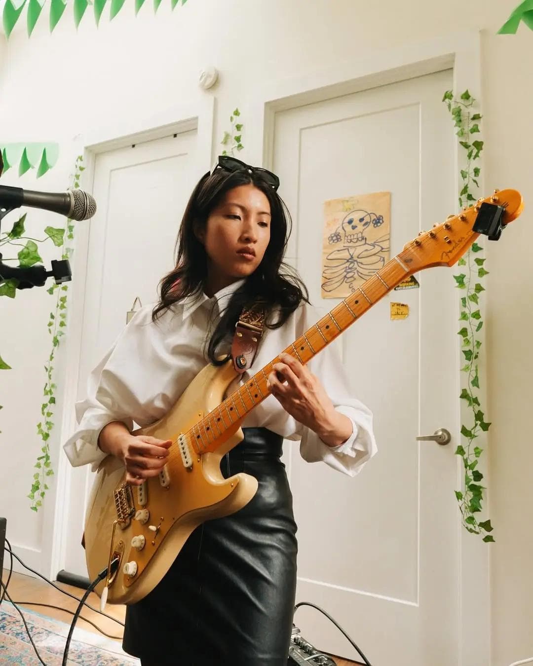 Instagram handle @ladiseoul. Electric guitarist performing indoors, wearing a white blouse and black skirt, with leafy decor on the walls.